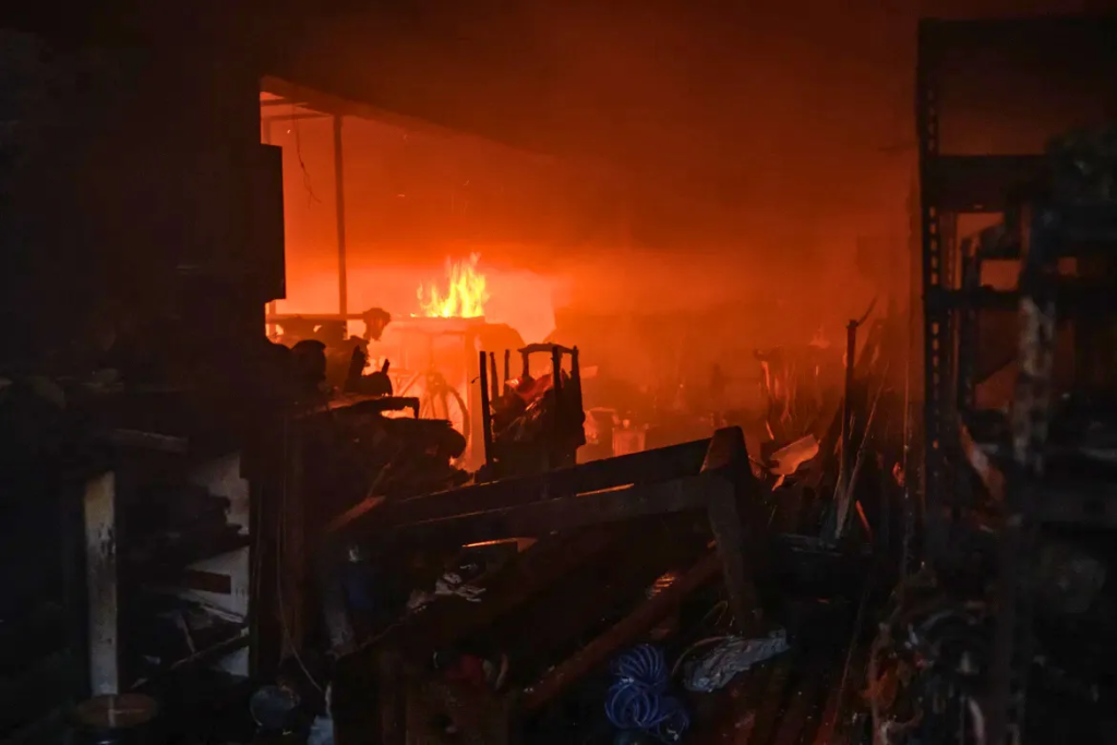 Destroyed home interior with charred debris and furniture silhouetted against orange glow from wildfire flames still burning outside in Greece