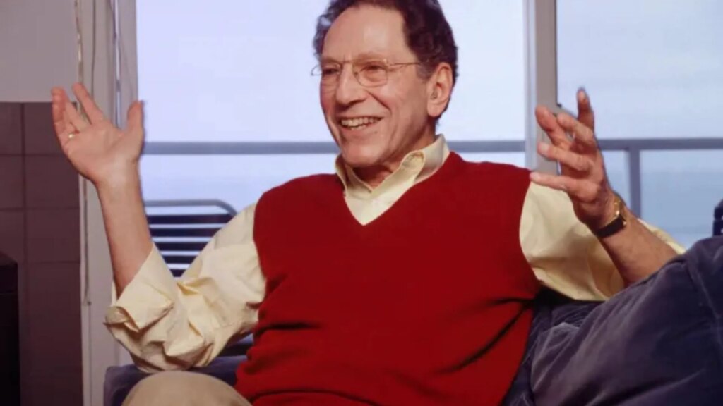 Tom Lehrer performing on stage, seated at a piano with a microphone, wearing a suit and glasses.
