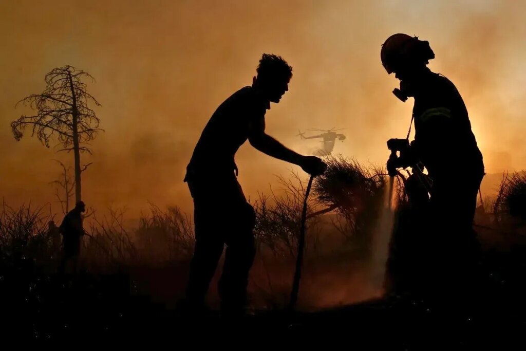 Silhouettes of two people working together with tools against orange smoky sky with helicopter in background during Greek wildfire emergency
