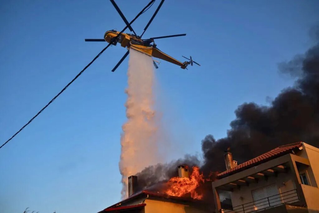 Yellow firefighting helicopter dropping water from above while house with red tile roof burns with orange flames and black smoke rising against blue sky in Greece